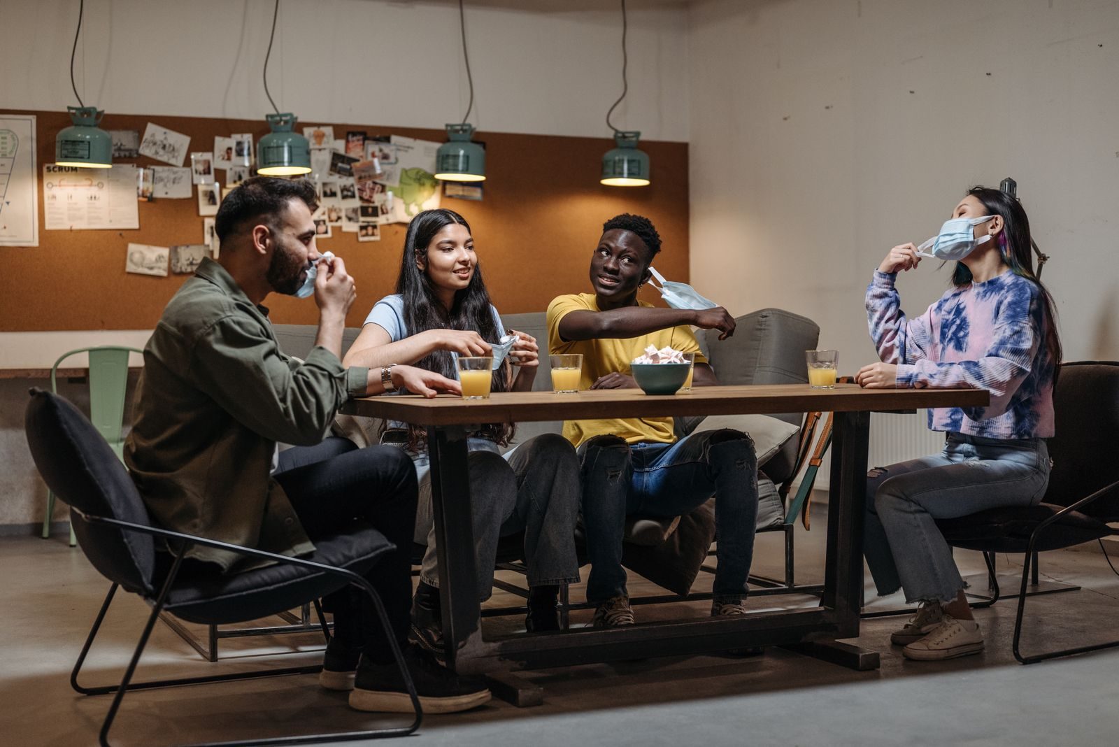 Grupo diverso de amigos à mesa com bebidas, socializando e tirando as máscaras em ambiente fechado