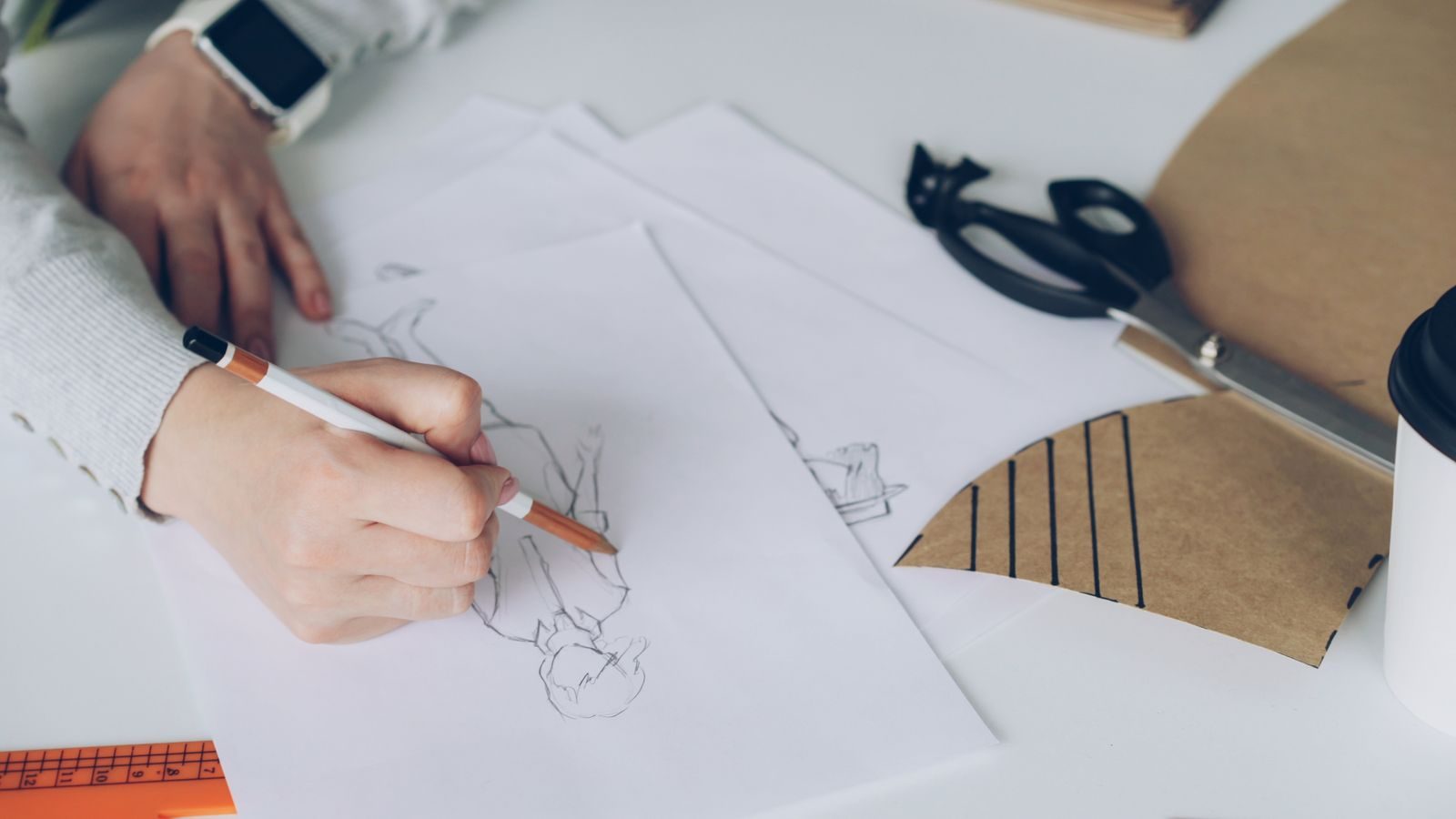 Mão de artista desenhando croquis de moda, com papéis e ferramentas à vista