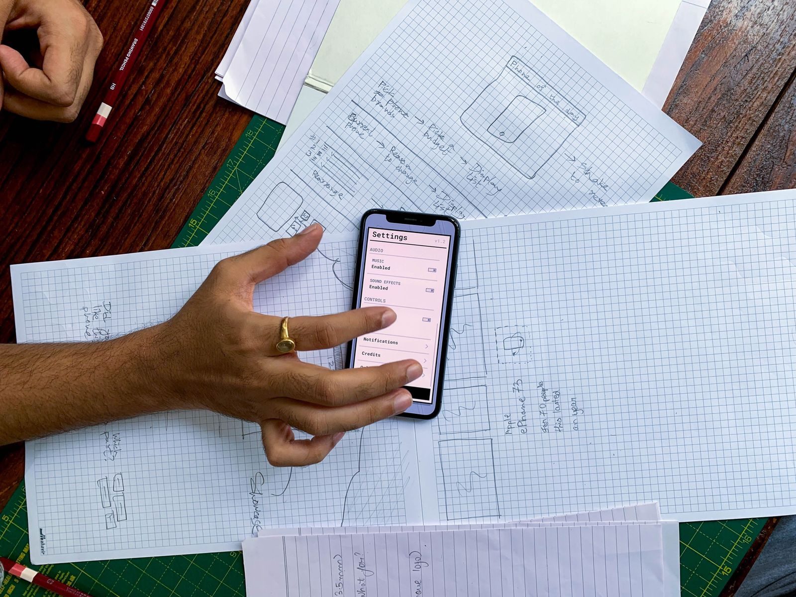 Mão segurando smartphone sobre esboços de design de app em papéis, vista de cima