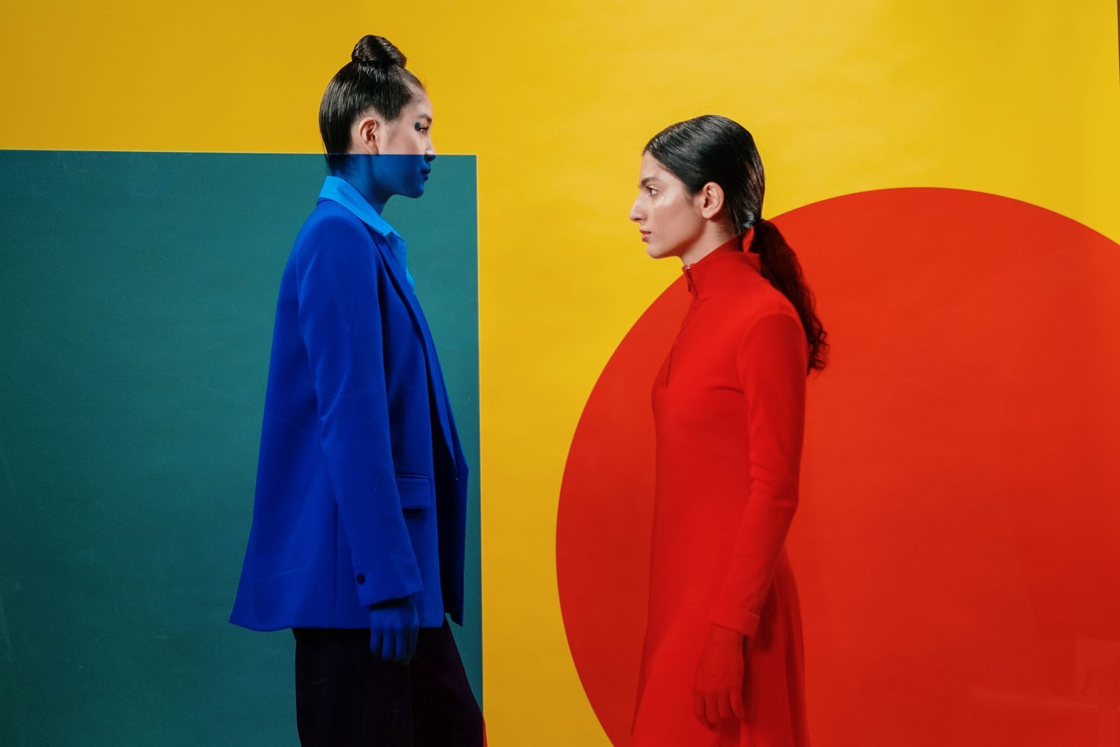 Retrato artístico de duas mulheres de vermelho e azul vibrantes diante de fundo geométrico