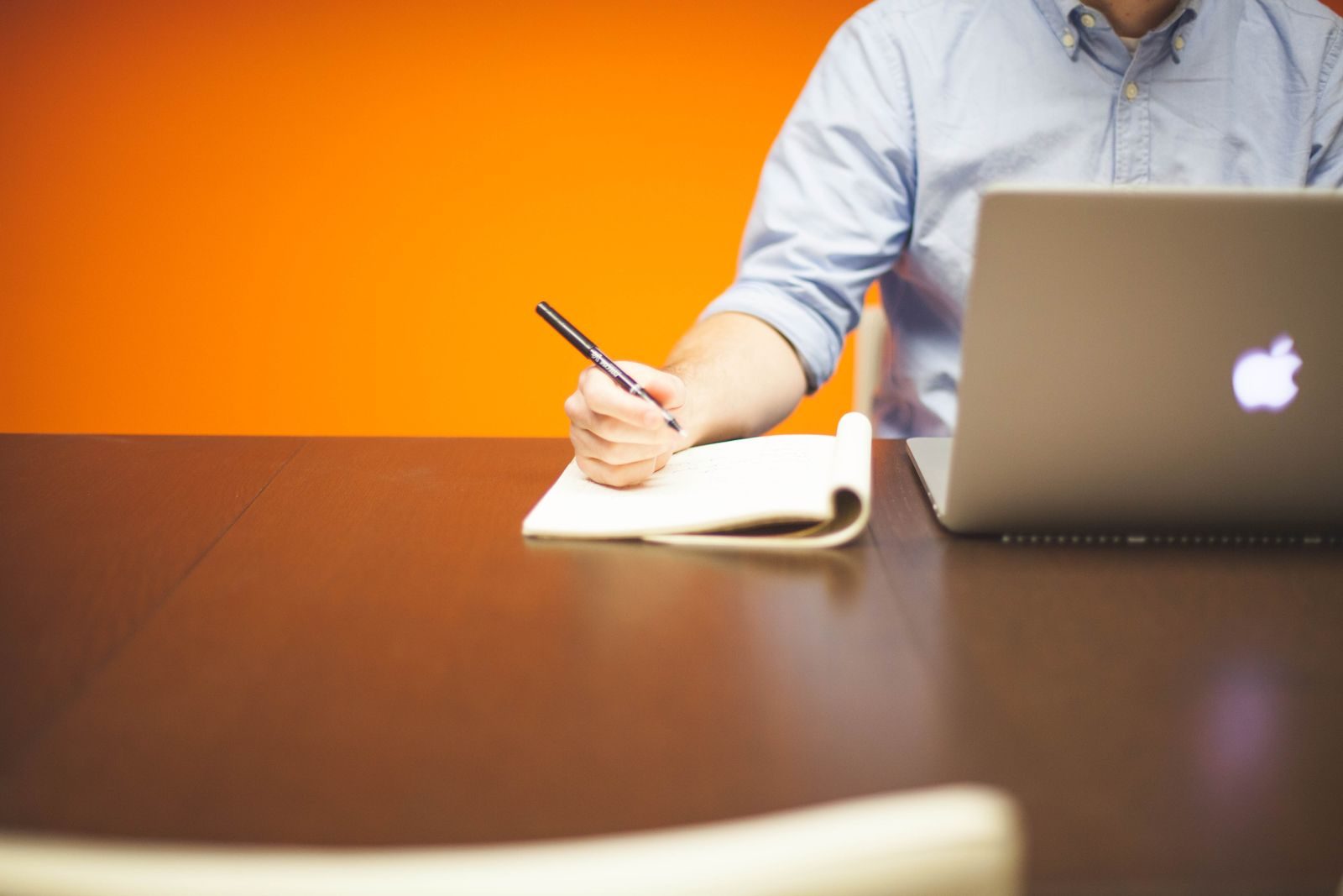 Adulto fazendo anotações em caderno ao lado de notebook em mesa de madeira com fundo laranja