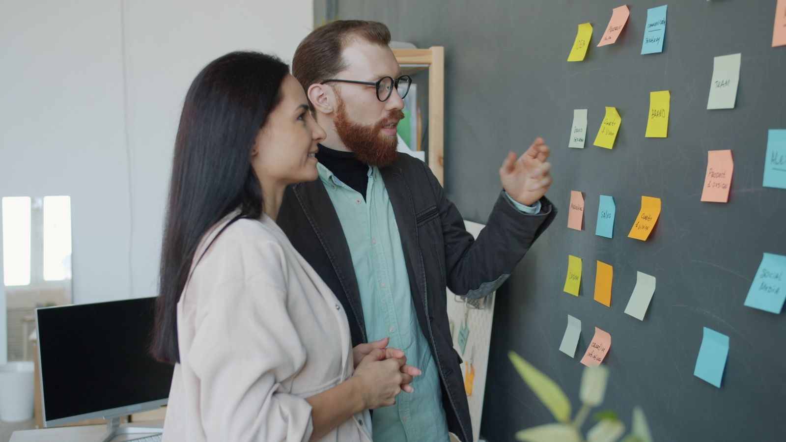 Homem e mulher fazendo brainstorming com post-its coloridos em um quadro-negro em escritório moderno