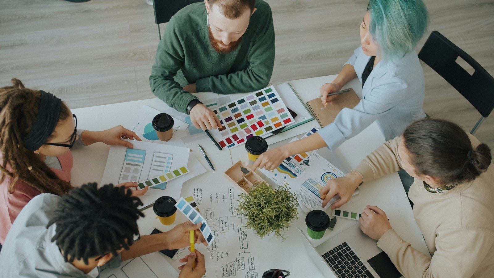Cinco pessoas colaborando em torno de uma mesa com amostras de cores, gráficos e xícaras de café