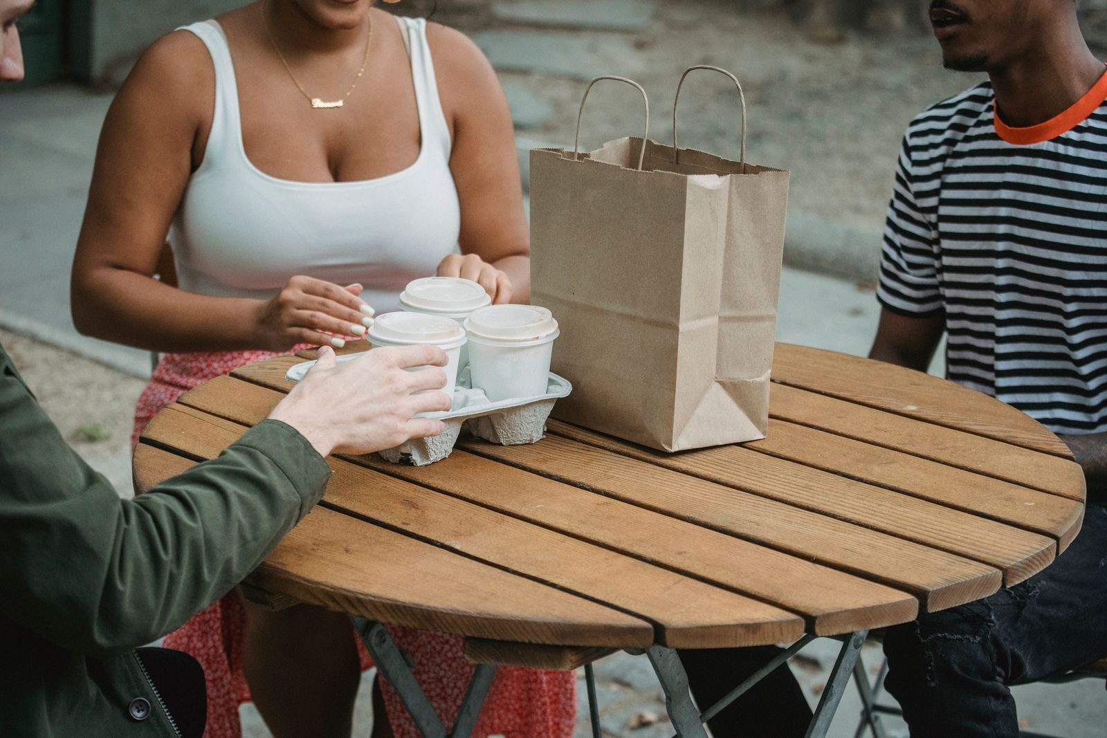 Vista do alto de jovens diversos em mesa redonda de madeira em café ao ar livre com bebidas para viagem e sacola de papel