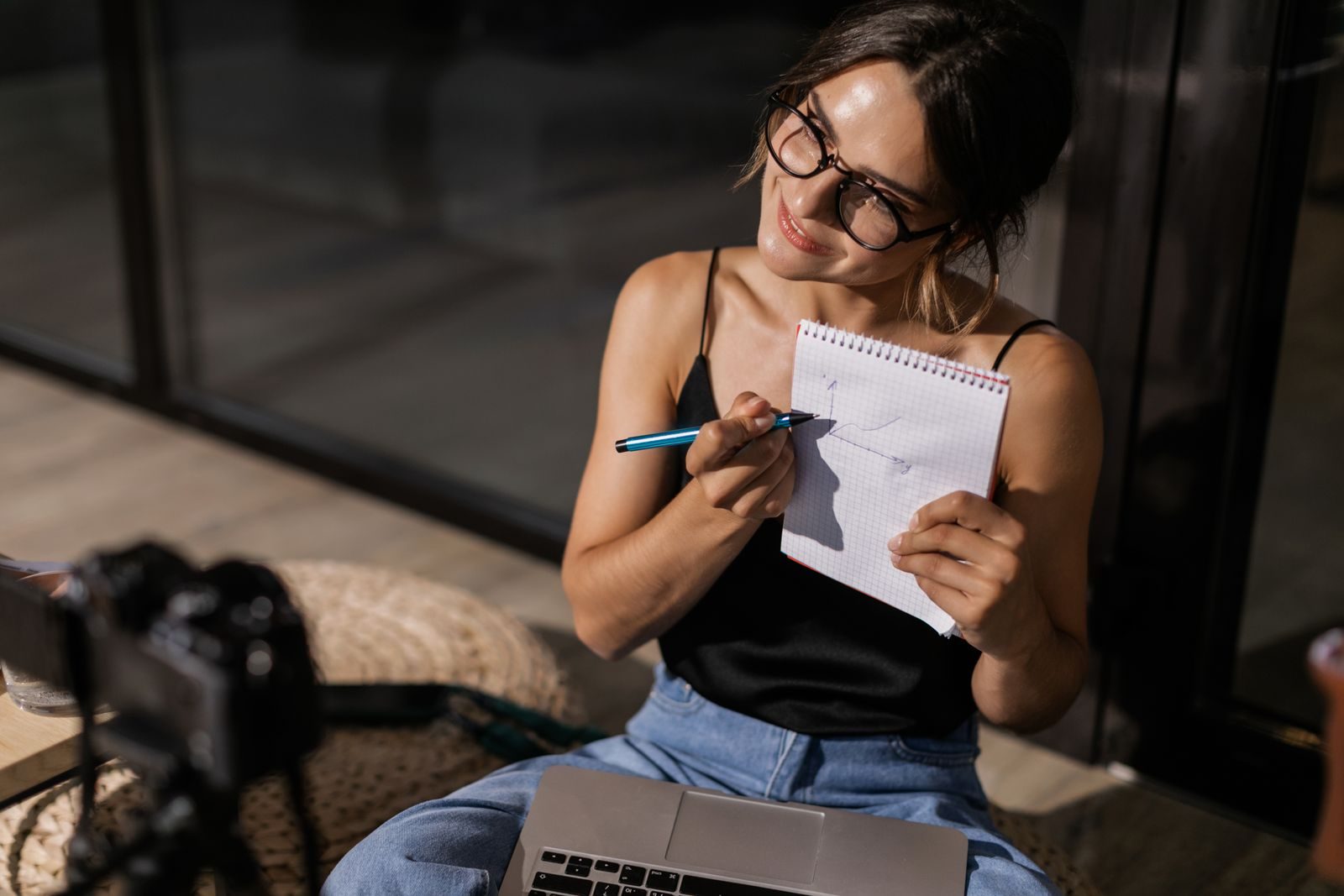 Mulher sorridente gravando vlog em ambiente interno com câmera, notebook e bloco de notas