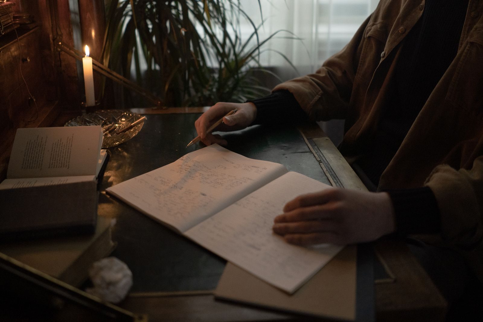Pessoa escrevendo em sala à meia-luz, cercada de livros abertos e velas, em clima intimista de estudo