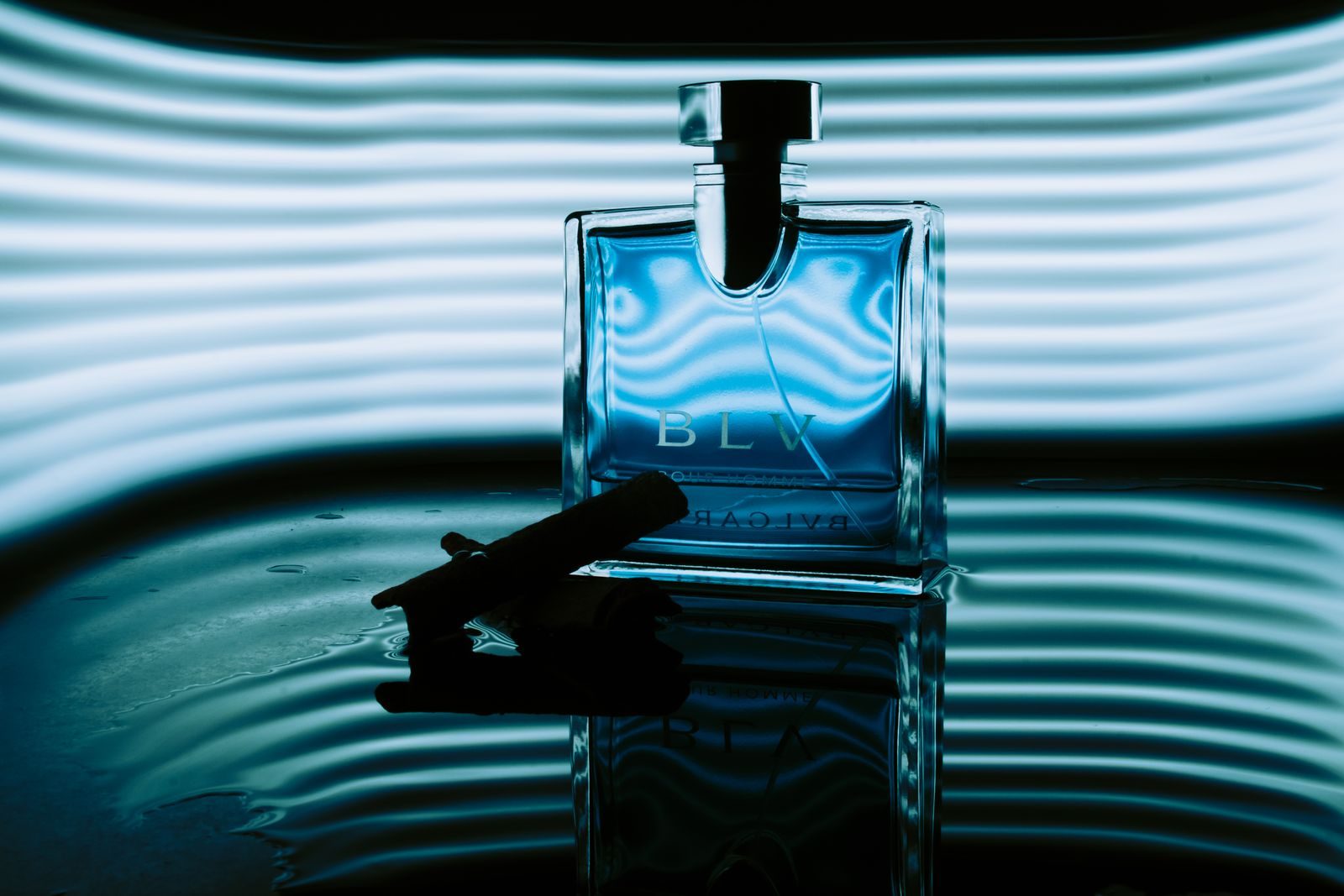 Close artístico de um frasco de perfume azul com iluminação ambiente e reflexo na água