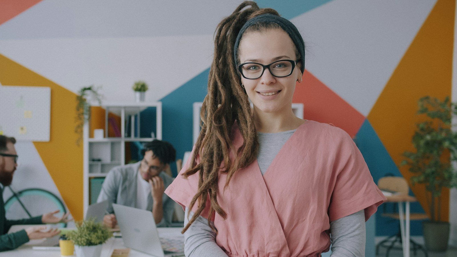 Mulher com dreads e óculos, de blusa rosa, em um escritório colorido