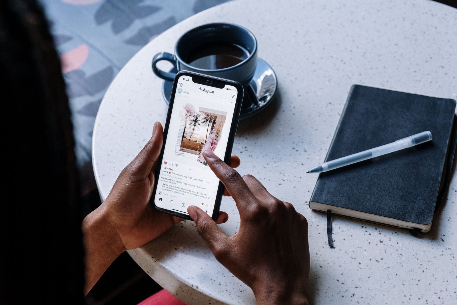 Mãos segurando smartphone em mesa de café com bebida e caderno, navegando em redes sociais