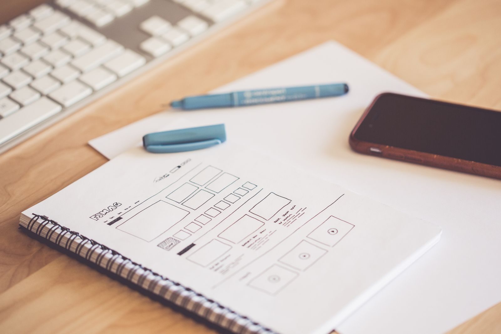 Mesa de madeira organizada com bloco de esboços com wireframes, smartphone e teclado