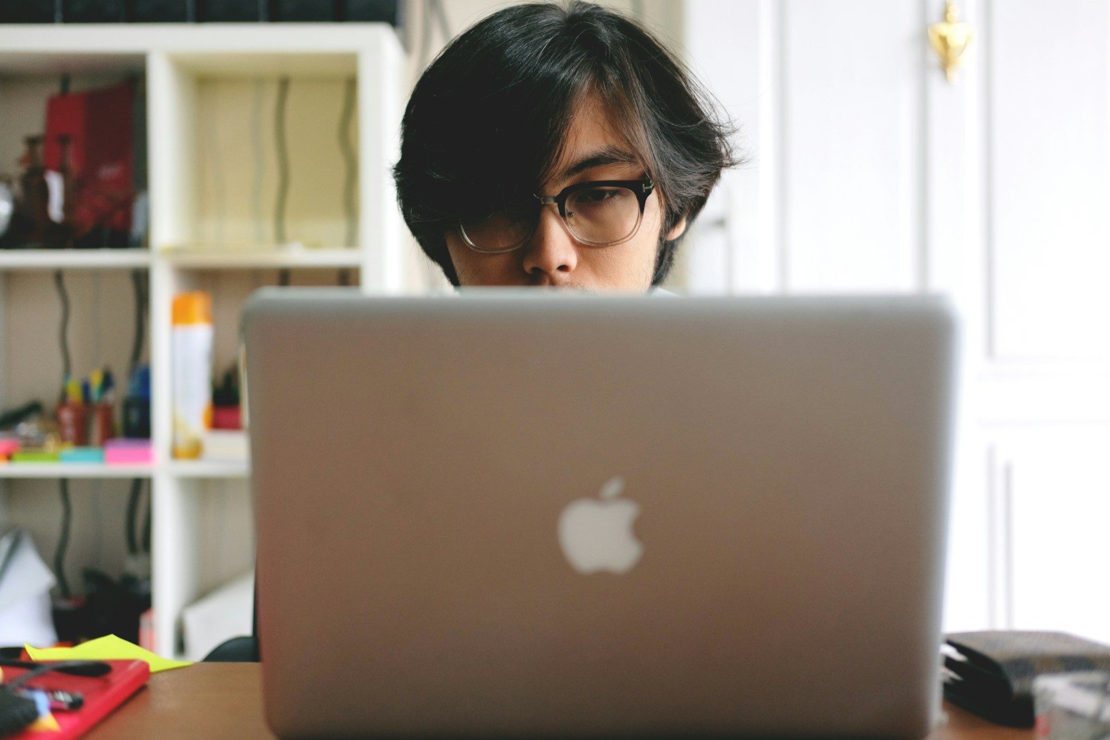Homem de óculos usando um MacBook prateado perto de uma estante de madeira em um cômodo