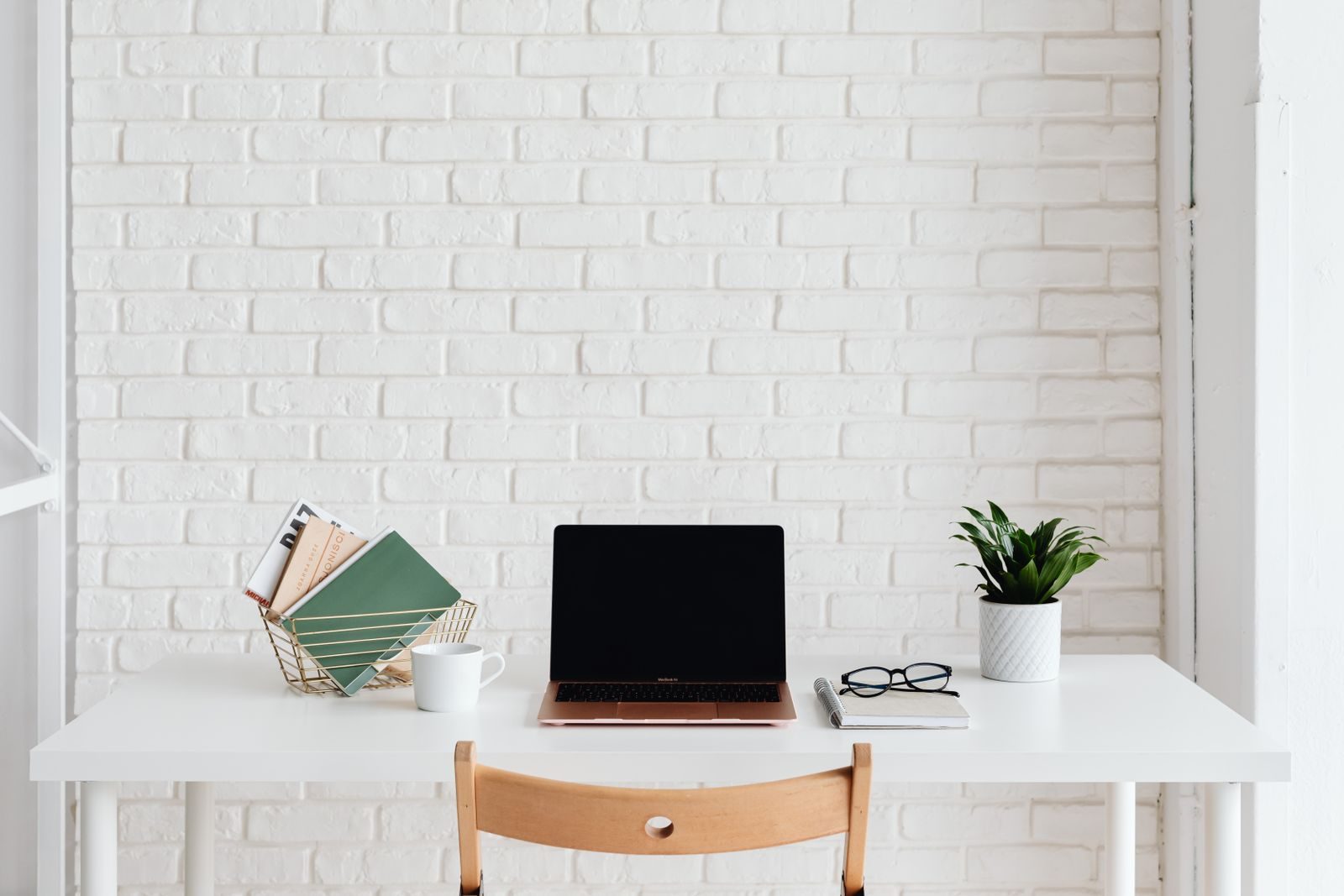 Espaço de trabalho branco e organizado com notebook, planta e livros diante de parede de tijolos