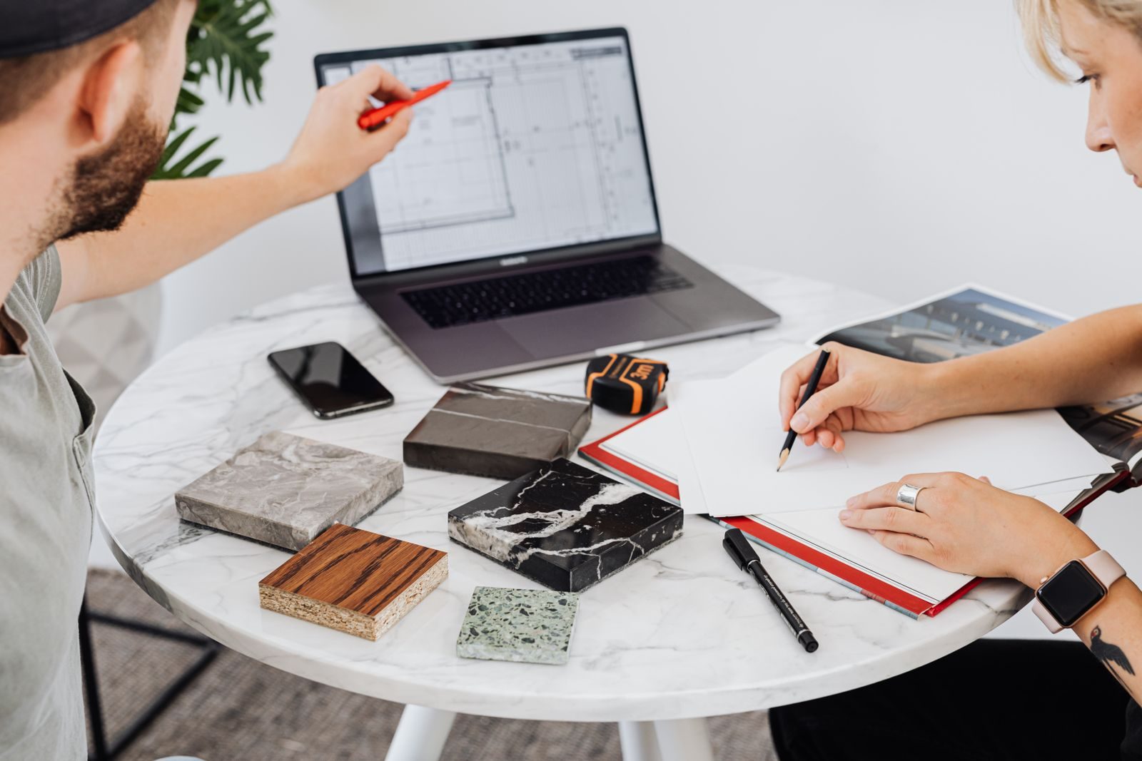 Dois designers colaborando com laptop e amostras de materiais em uma mesa de mármore