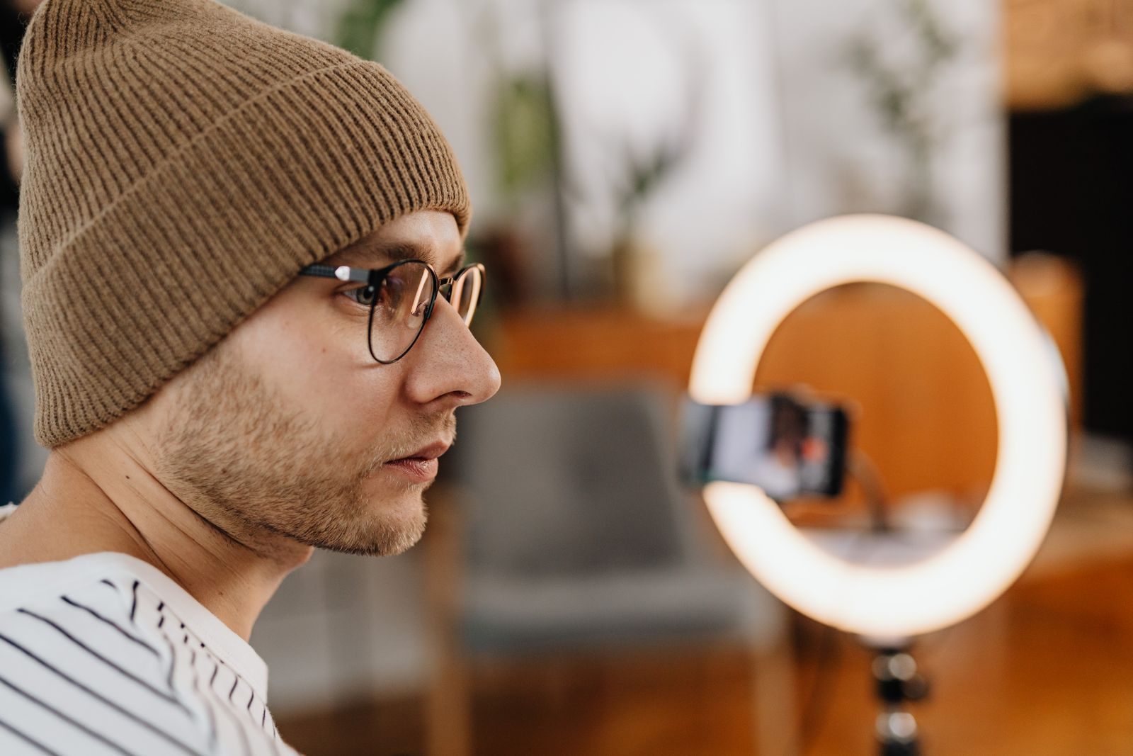 Homem de óculos e gorro de tricô gravando vídeo em casa com smartphone e ring light