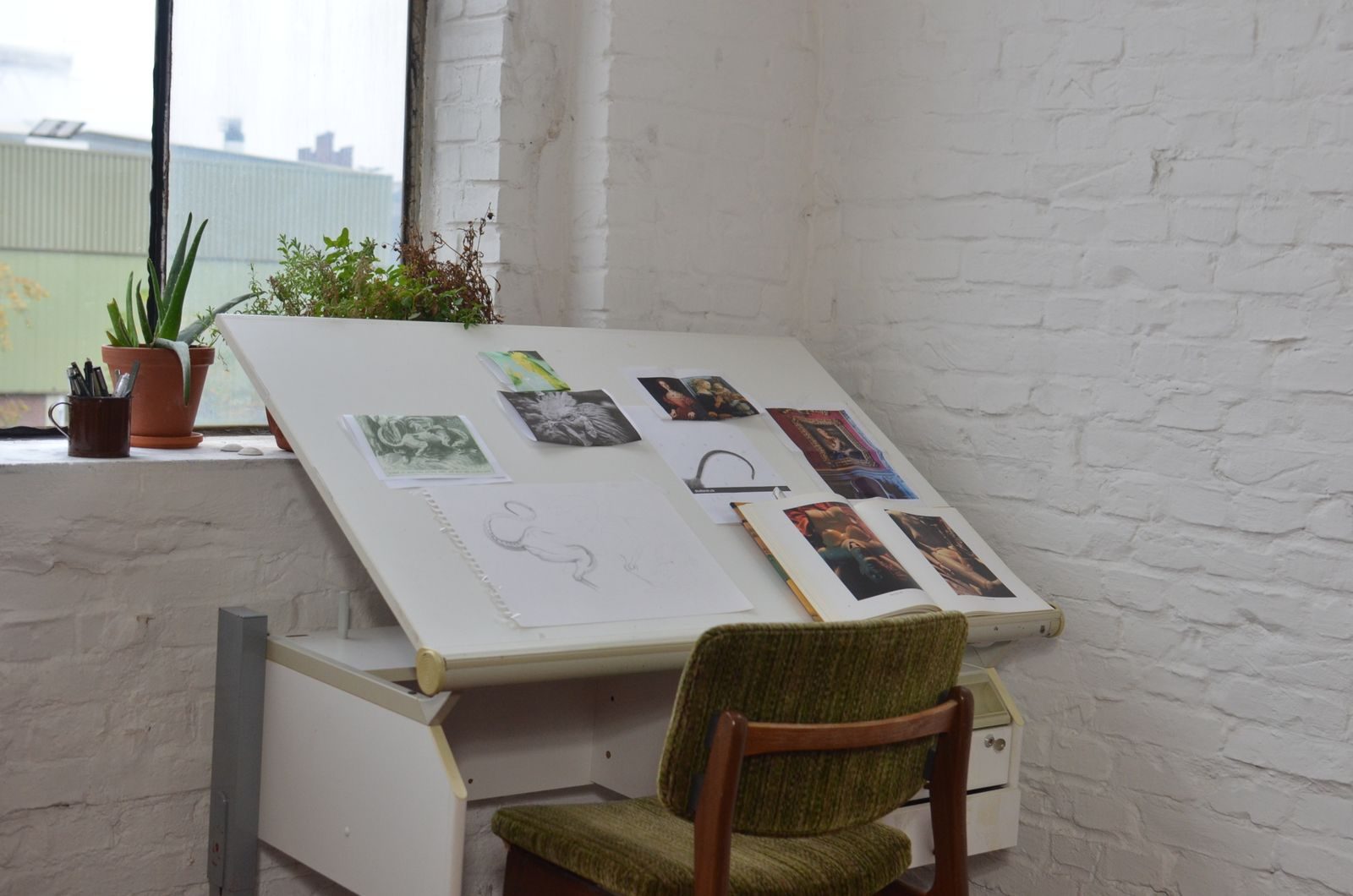 Ateliê de arte com desenhos e esboços na mesa, cadeira, vaso de planta e materiais de papelaria na janela