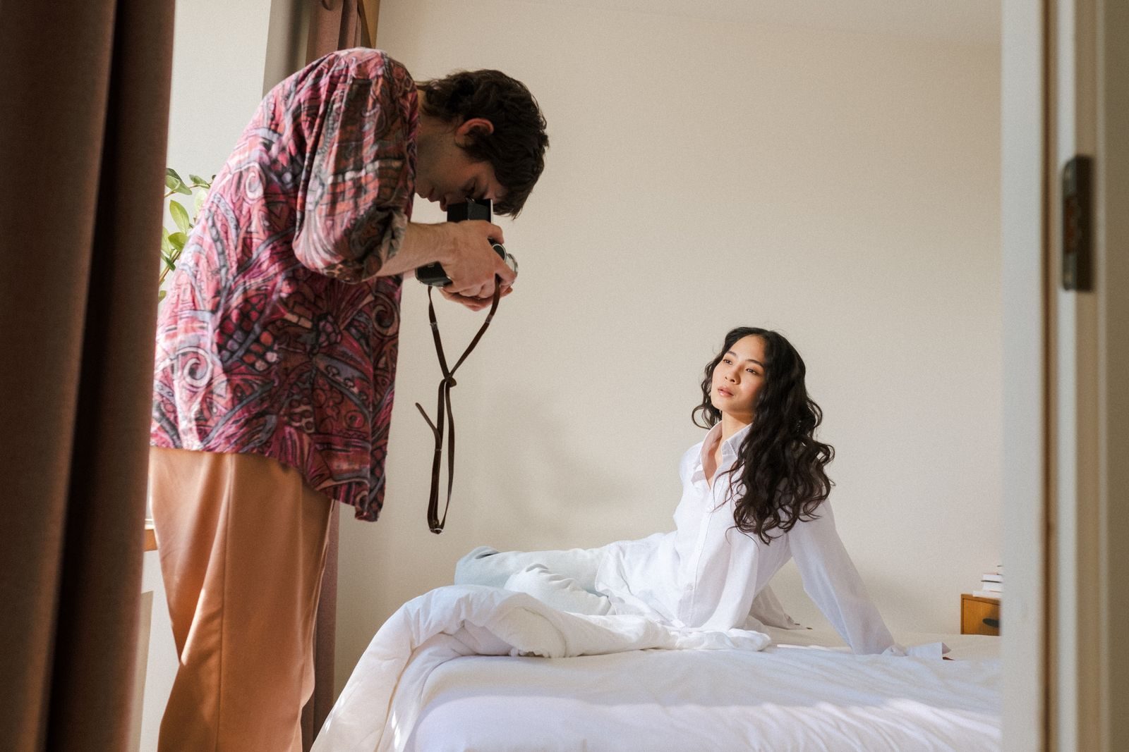 Modelo posando na cama enquanto fotógrafo usa câmera analógica em ensaio criativo