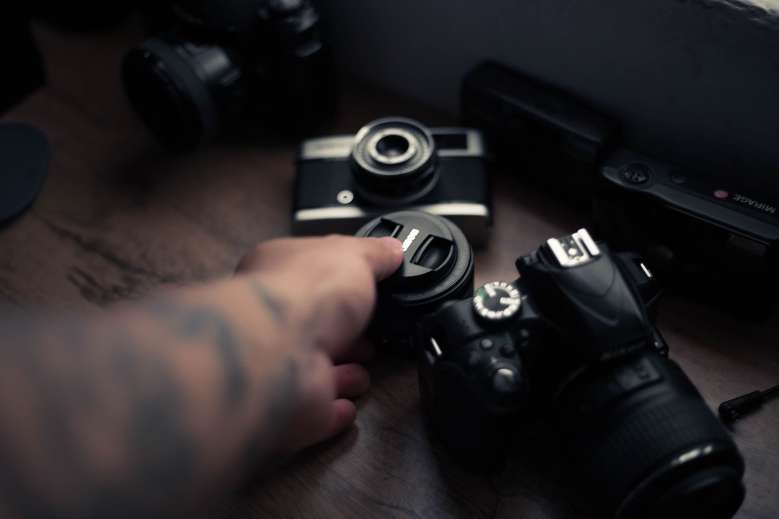 Mão segurando lente de câmera entre equipamentos fotográficos sobre mesa de madeira