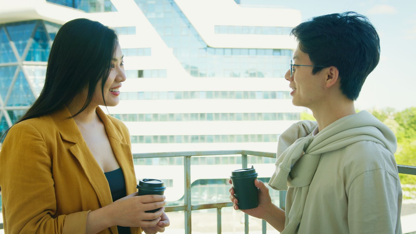 Dois amigos conversando e tomando café em uma varanda urbana com prédio moderno ao fundo
