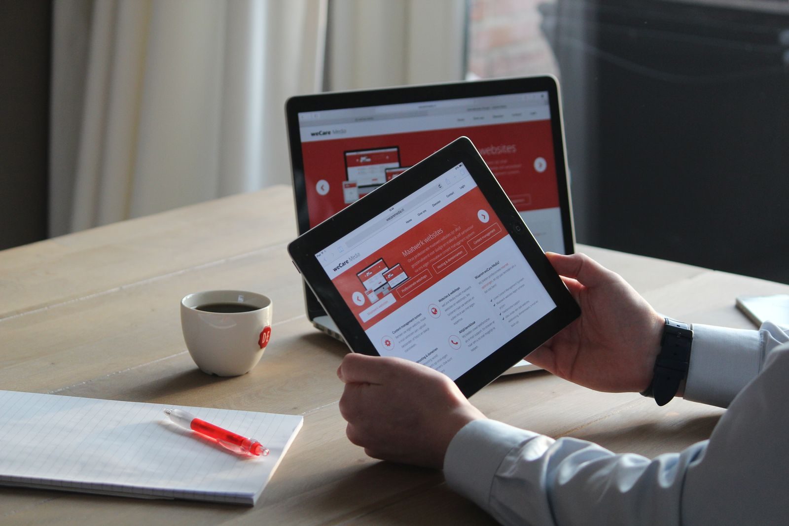 Profissional usando tablet e notebook com bebida quente, revisando conteúdo digital