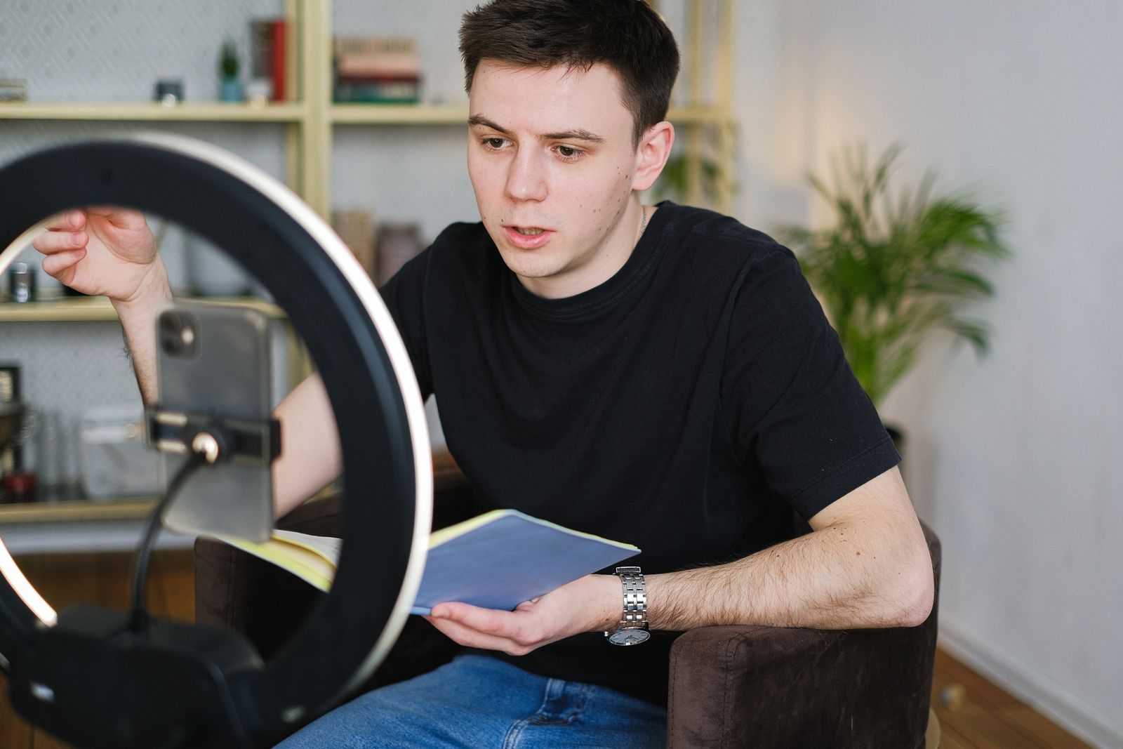 Homem gravando vídeo em ambiente interno com smartphone e ring light, segurando um caderno
