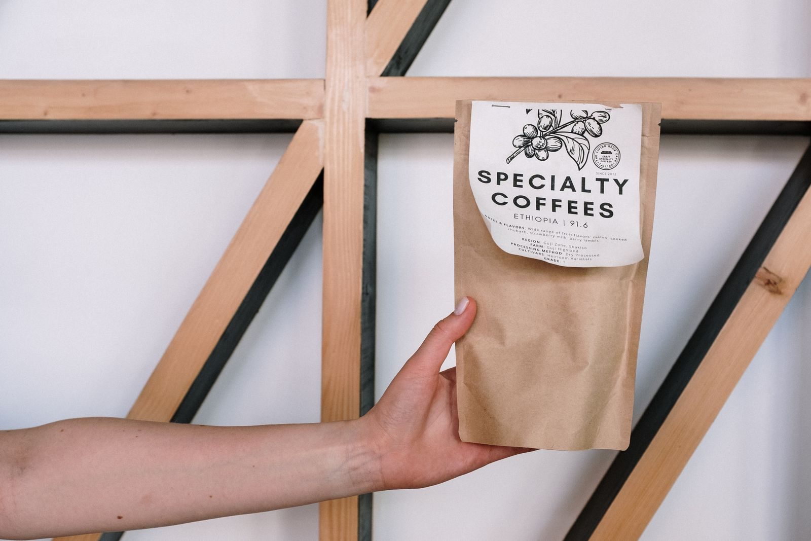 Mão segurando um pacote de café especial contra fundo minimalista com vigas de madeira