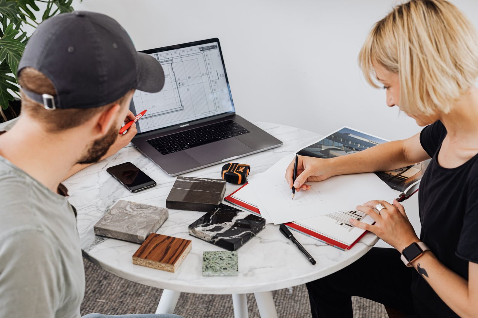 Dois designers colaborando em um projeto com laptop e materiais de desenho em uma mesa redonda