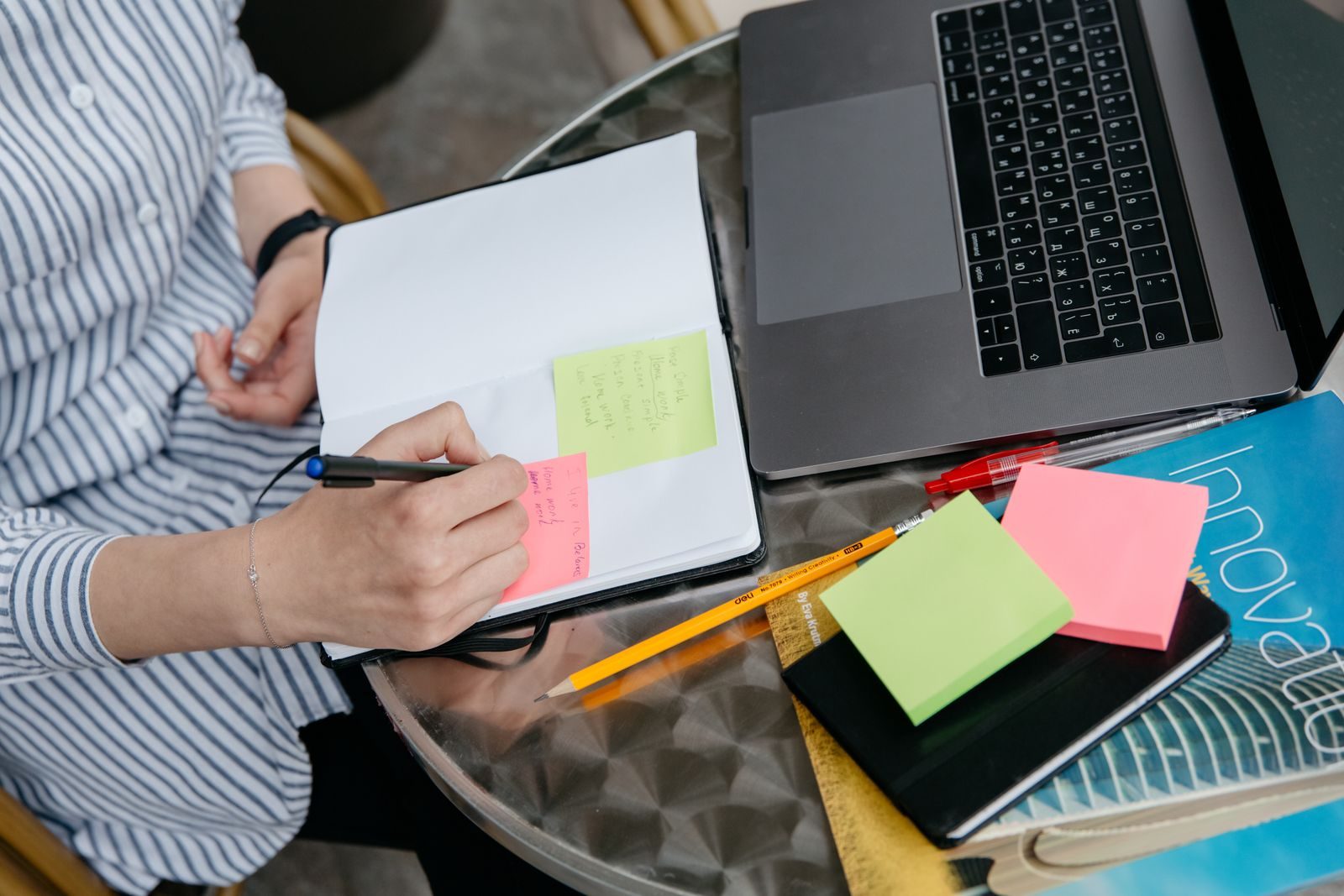 Pessoa escrevendo em caderno com notebook e post-its na mesa, em um espaço de trabalho criativo e organizado