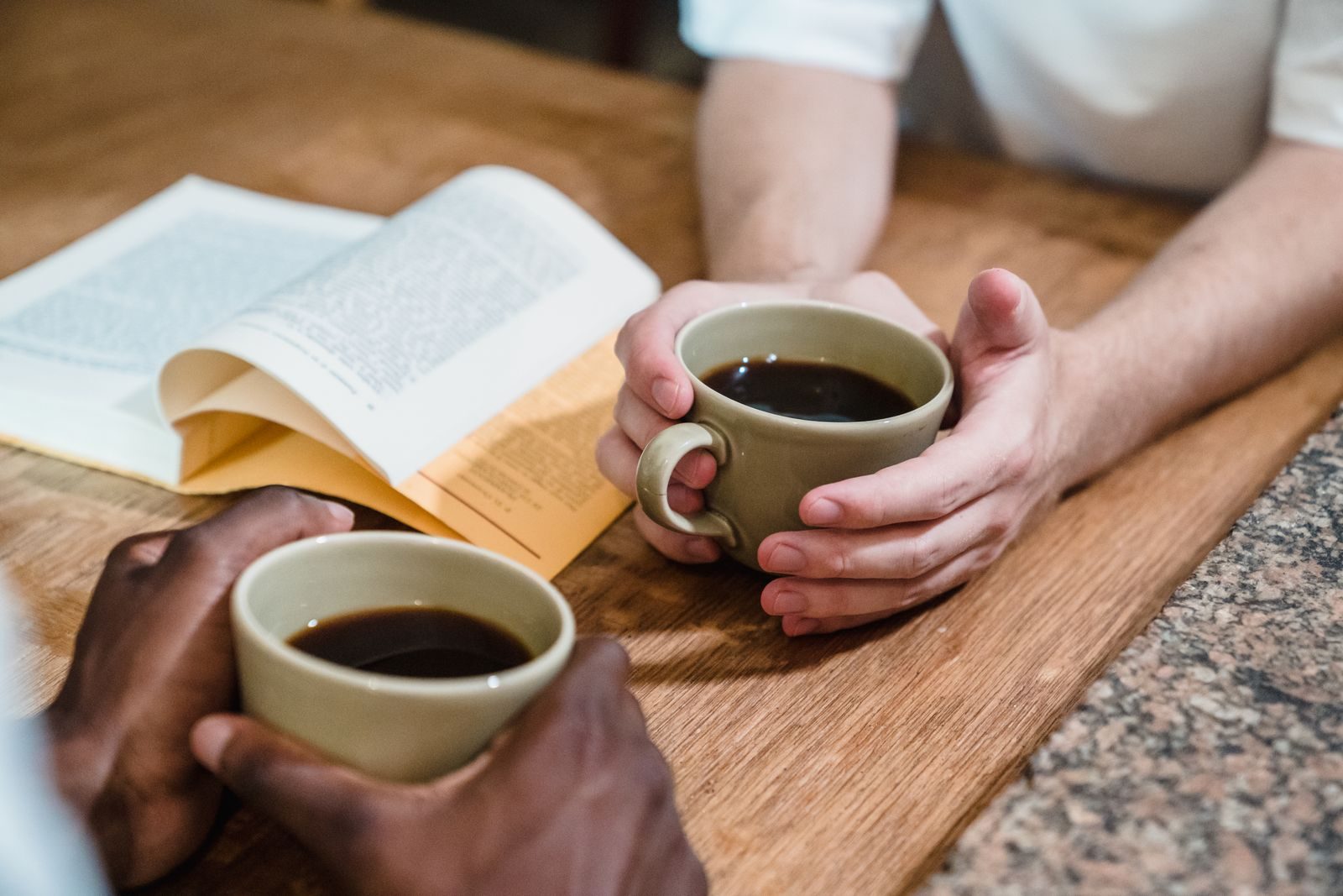 Duas pessoas segurando xícaras de café e lendo um livro em uma mesa de madeira