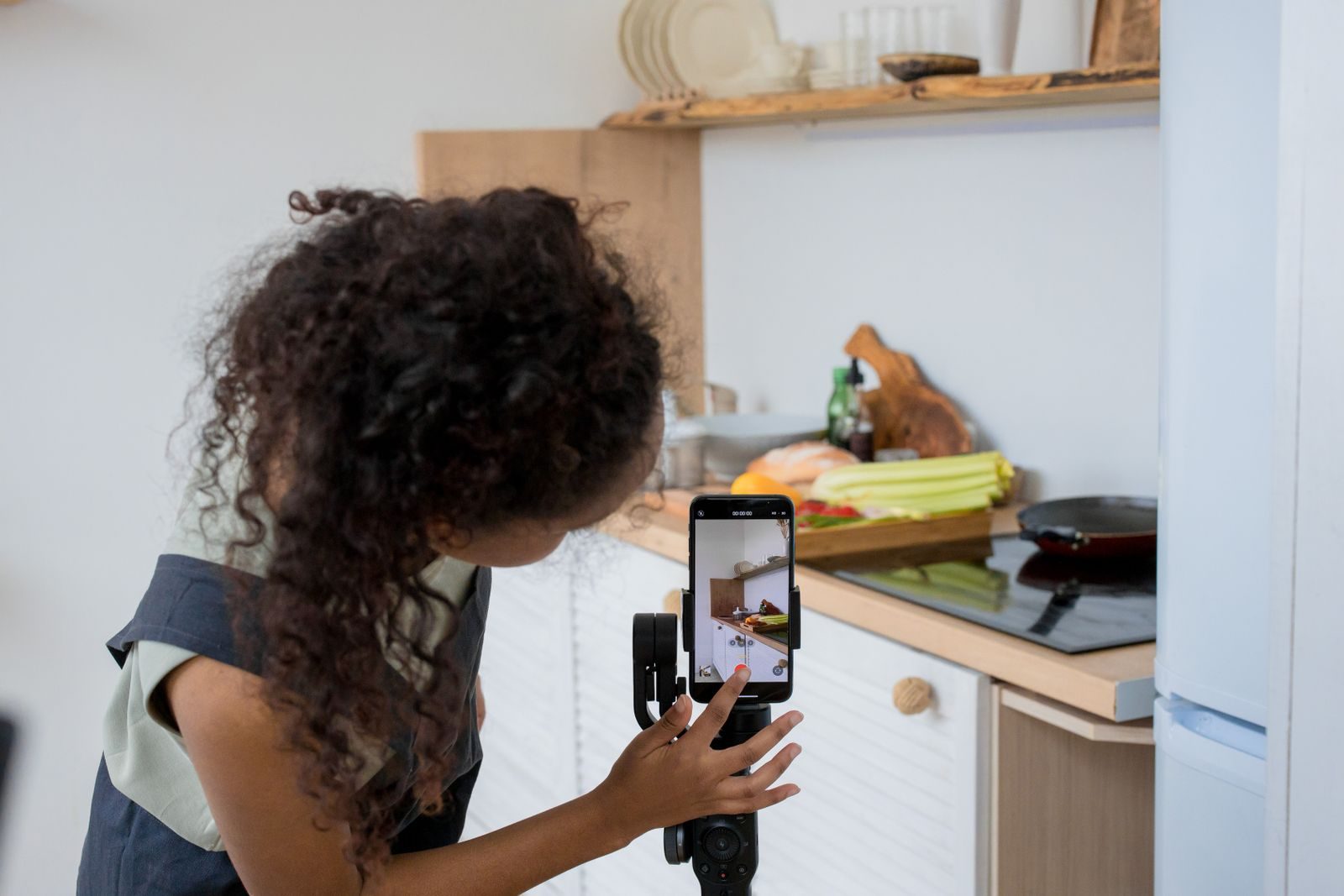 Mulher de cabelo cacheado gravando vídeo de culinária com estabilizador de celular em cozinha moderna