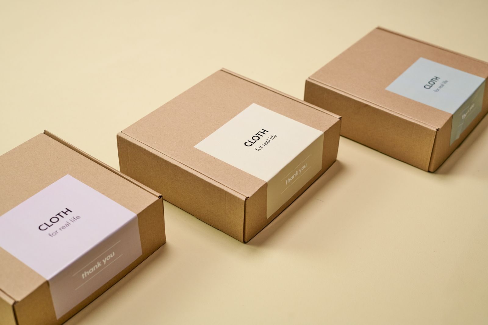 Três embalagens minimalistas de papelão com o rótulo 'CLOTH' em fundo amarelo