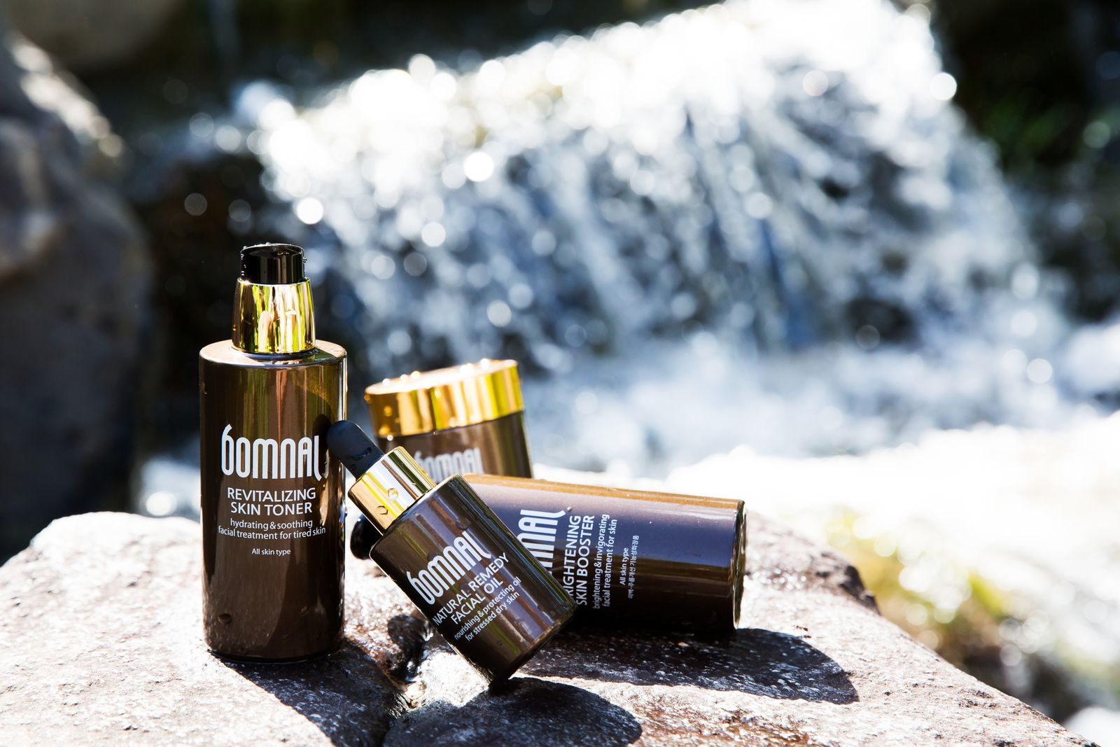 Close de produtos de skincare Bomnal sobre pedras perto de uma cachoeira, com embalagem luxuosa