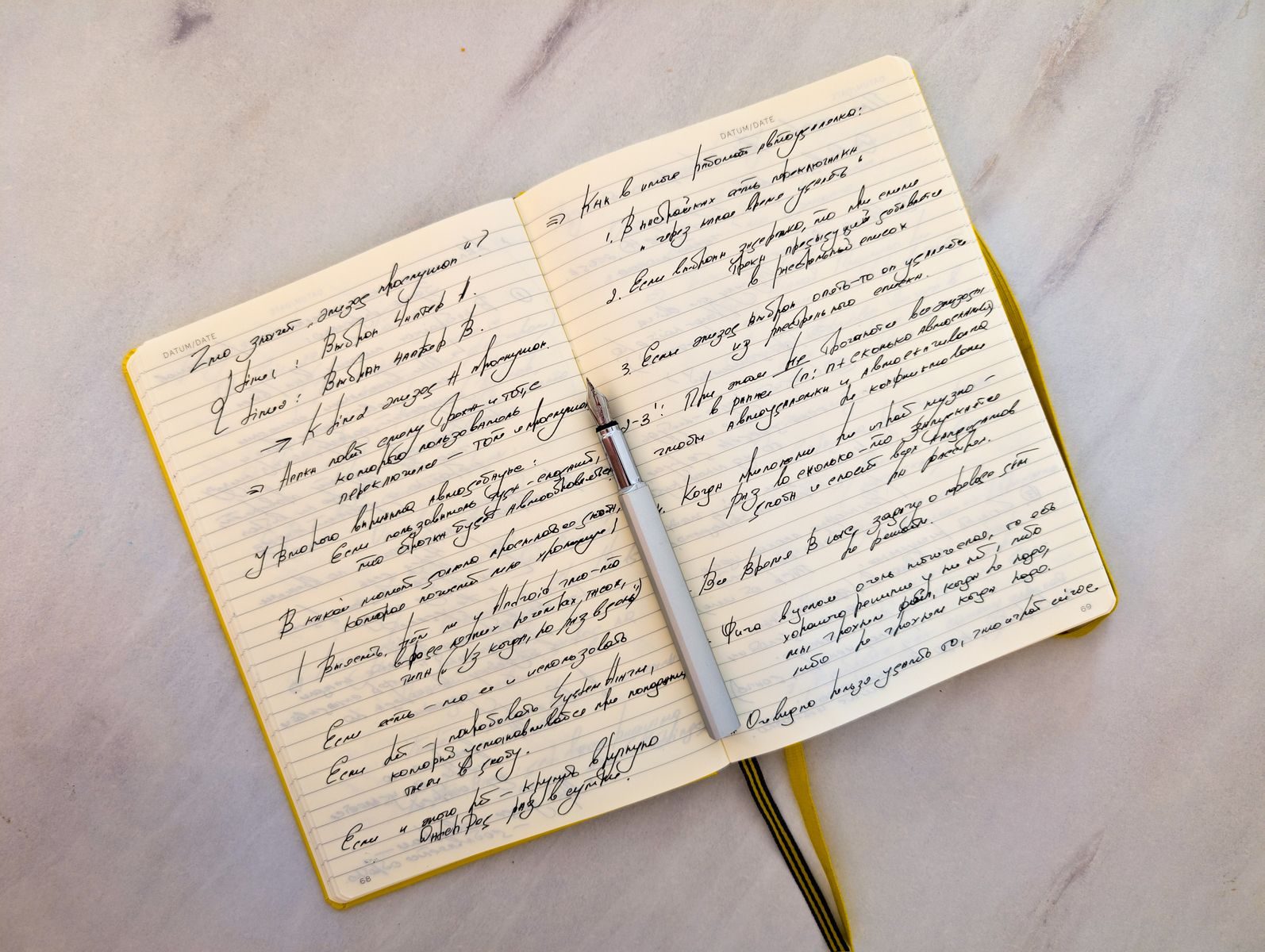 Caderno aberto com anotações em letra cursiva sobre superfície de mármore e caneta apoiada nas páginas