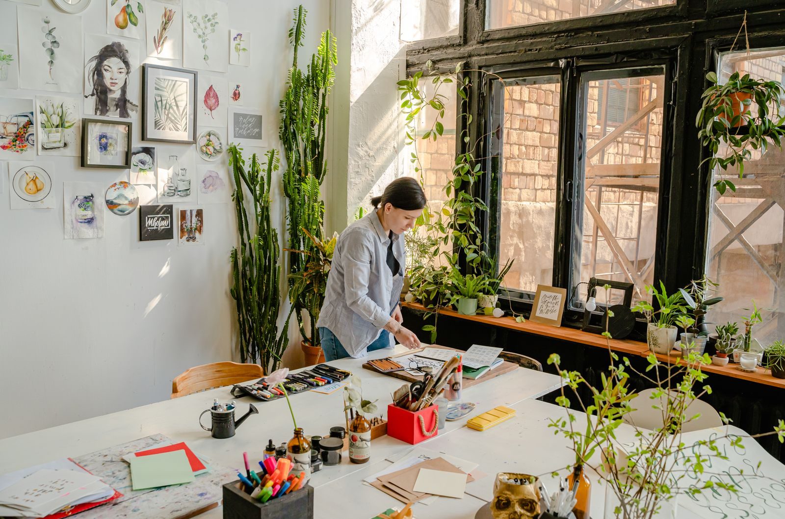 Designer desenhando na mesa com canetinhas e papéis, perto de parede com obras de arte e plantas na janela