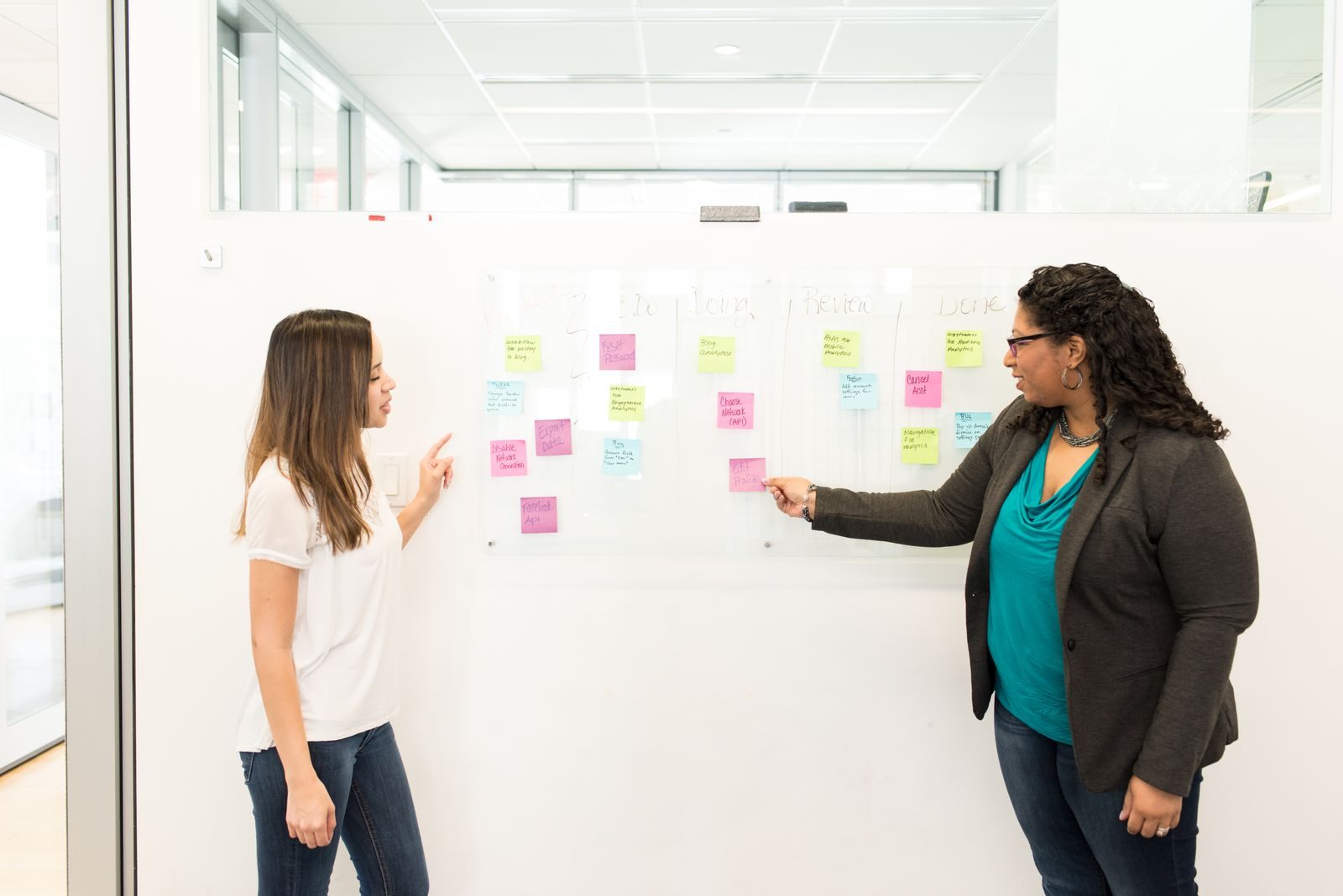 Duas mulheres conversando em um escritório com post-its em um quadro branco, em clima de trabalho em equipe