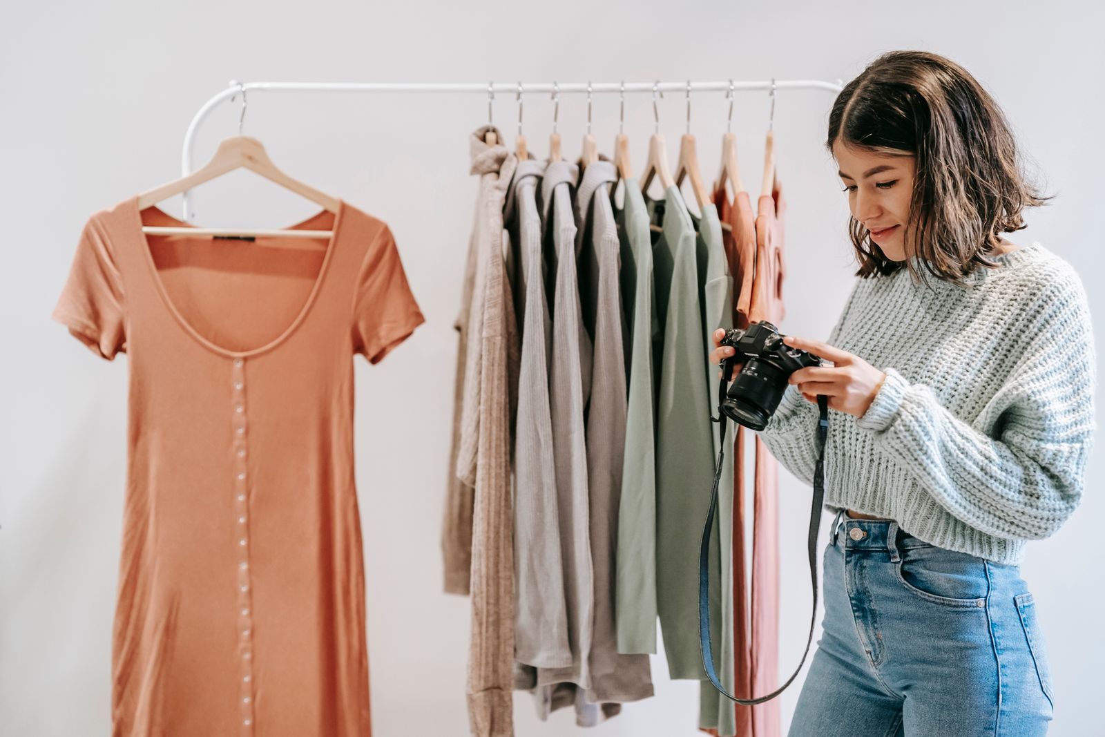 Jovem latino-americana sorrindo com câmera profissional diante de arara com roupas variadas