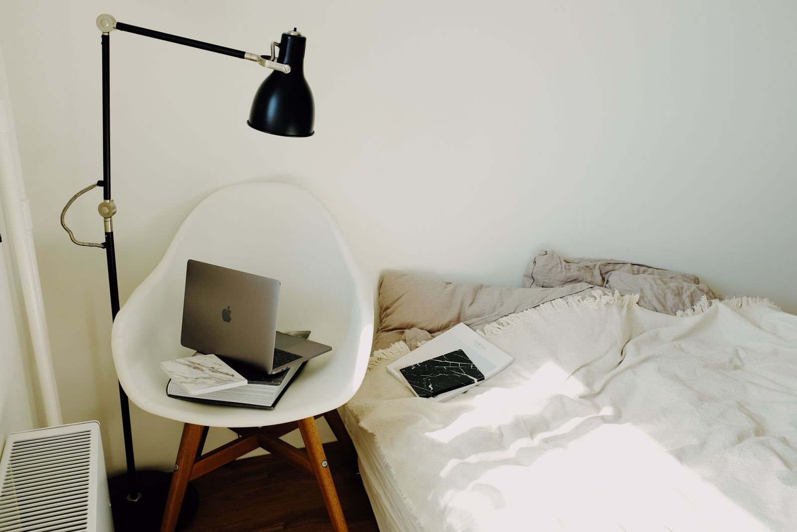 Vista do alto de notebook sobre livro preto em poltrona branca, sob luminária, perto da cama em quarto claro e moderno