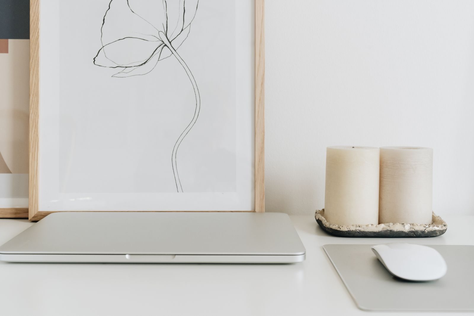 Home office minimalista com notebook fechado, quadro emoldurado e velas em mesa branca